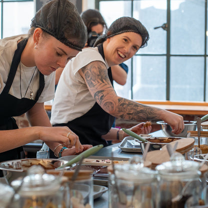 Two women wearing black aprons and headbands work at a food prep station with glass jars and ingredients in a bright kitchen or bakery setting.