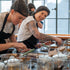 Two women wearing black aprons and headbands work at a food prep station with glass jars and ingredients in a bright kitchen or bakery setting.