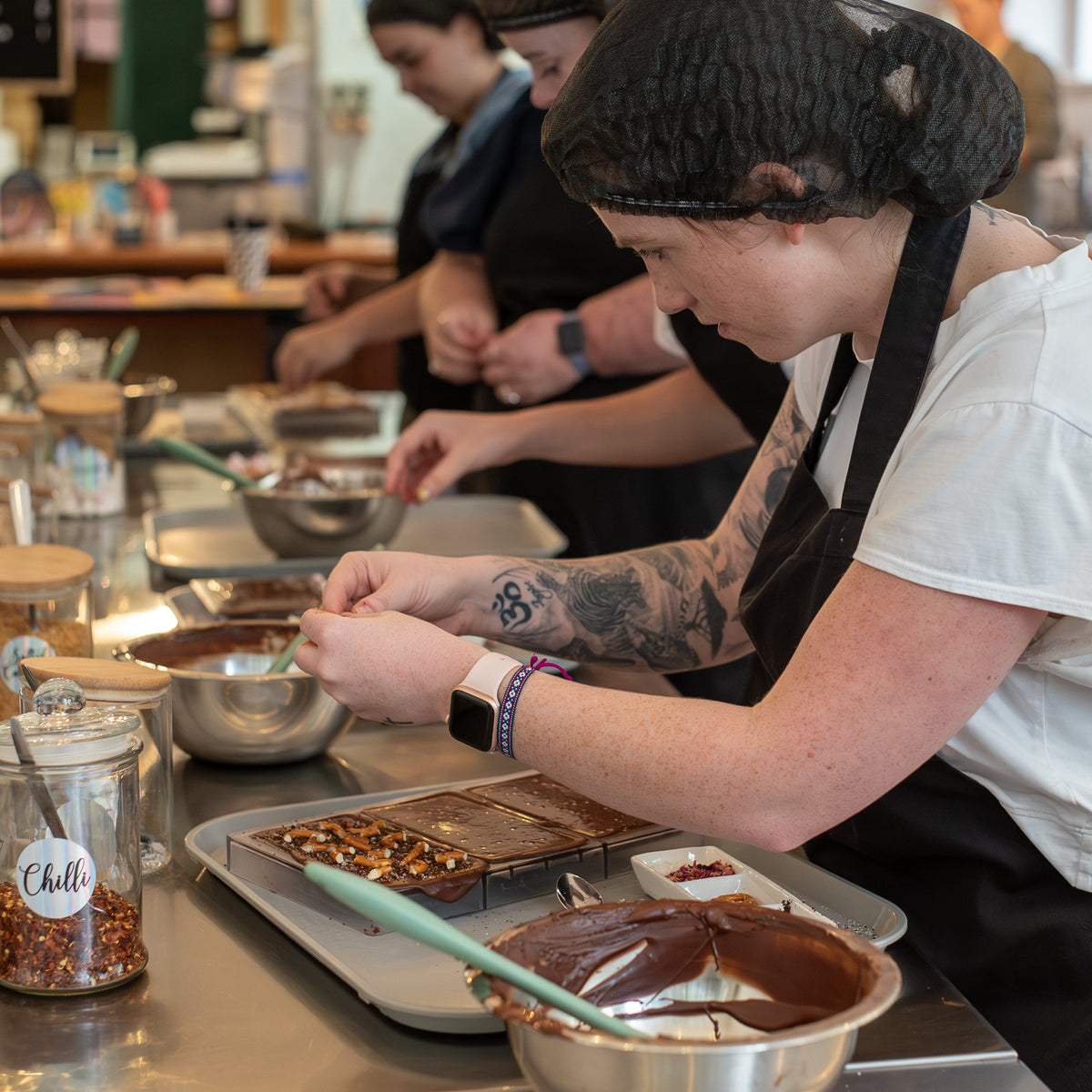A woman in a white t-shirt and black apron works at a chocolate station, dipping a chocolate bar into melted chocolate. Other staff members and glass jars are visible in the background of a cafe or bakery setting.