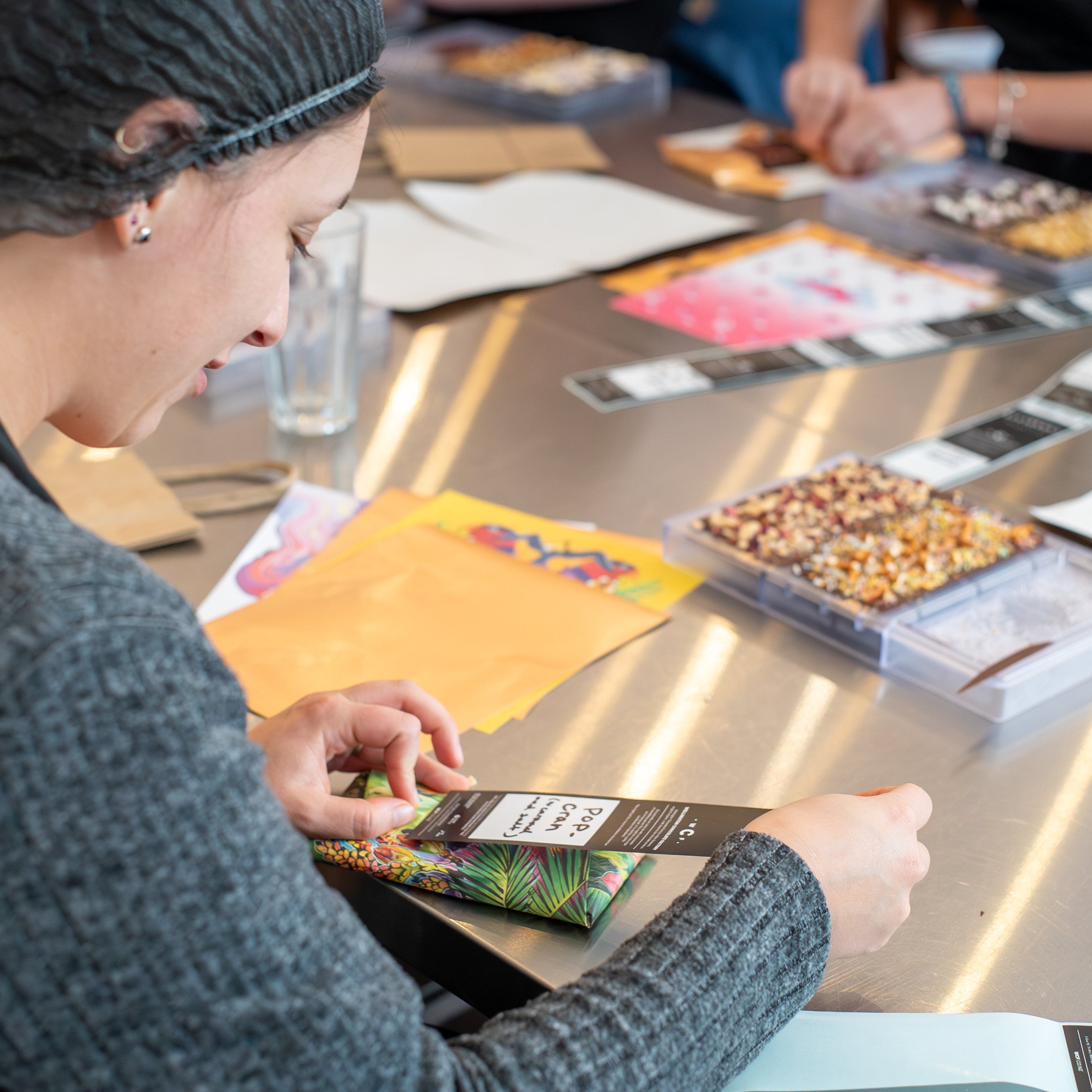 A person wearing a black hair net and gray sweater sits at a table, wrapping a colorful packaged item. The table is covered with papers, small trays of mixed materials, and other crafting or packaging supplies in a bright indoor workspace.