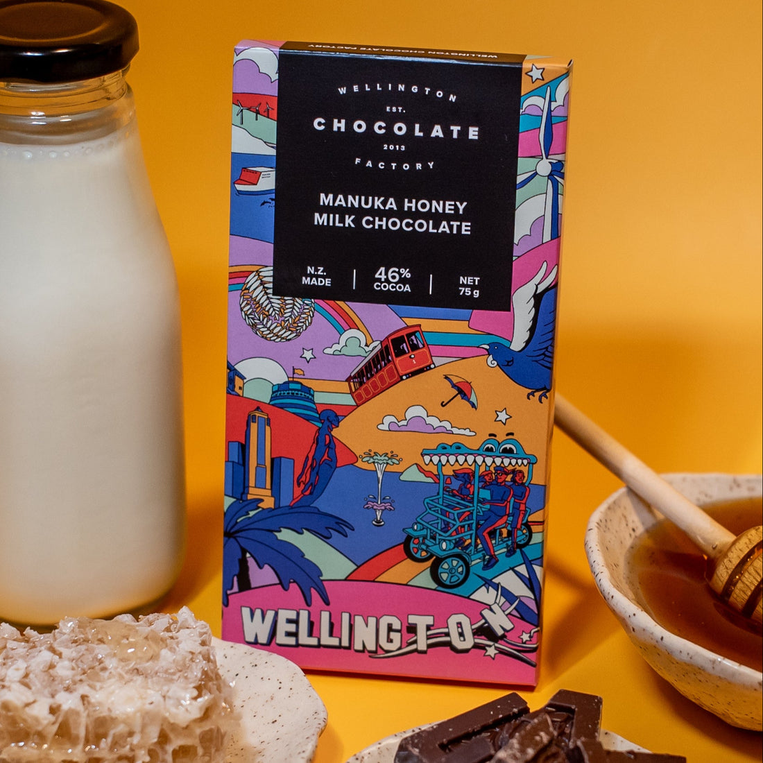Manuka Honey Milk Chocolate bar from The Wellington Chocolate Factory with honeycomb, milk, and a honey dipper on an orange background.